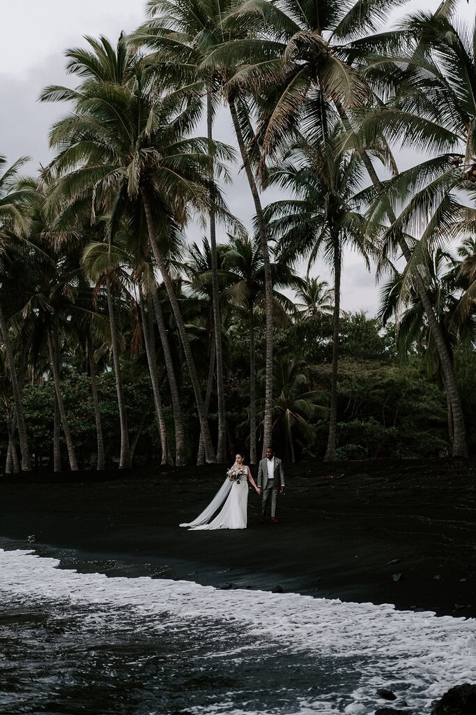 newlyweds holding hands walking on a black sand beach with palm trees in the background.