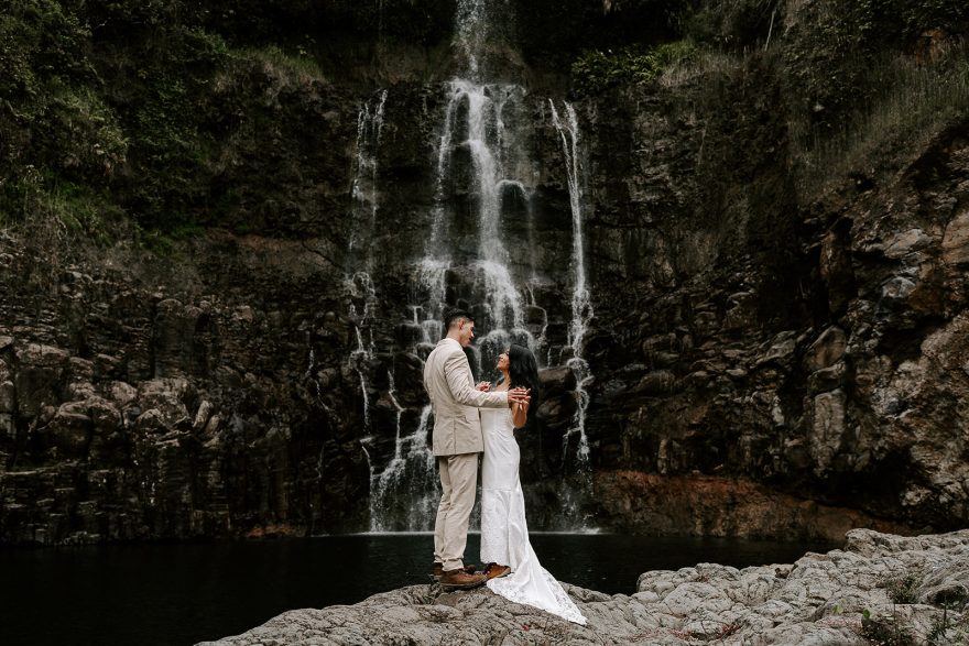 Newlyweds holding hands by a large waterfall as they elope in Hawaii.