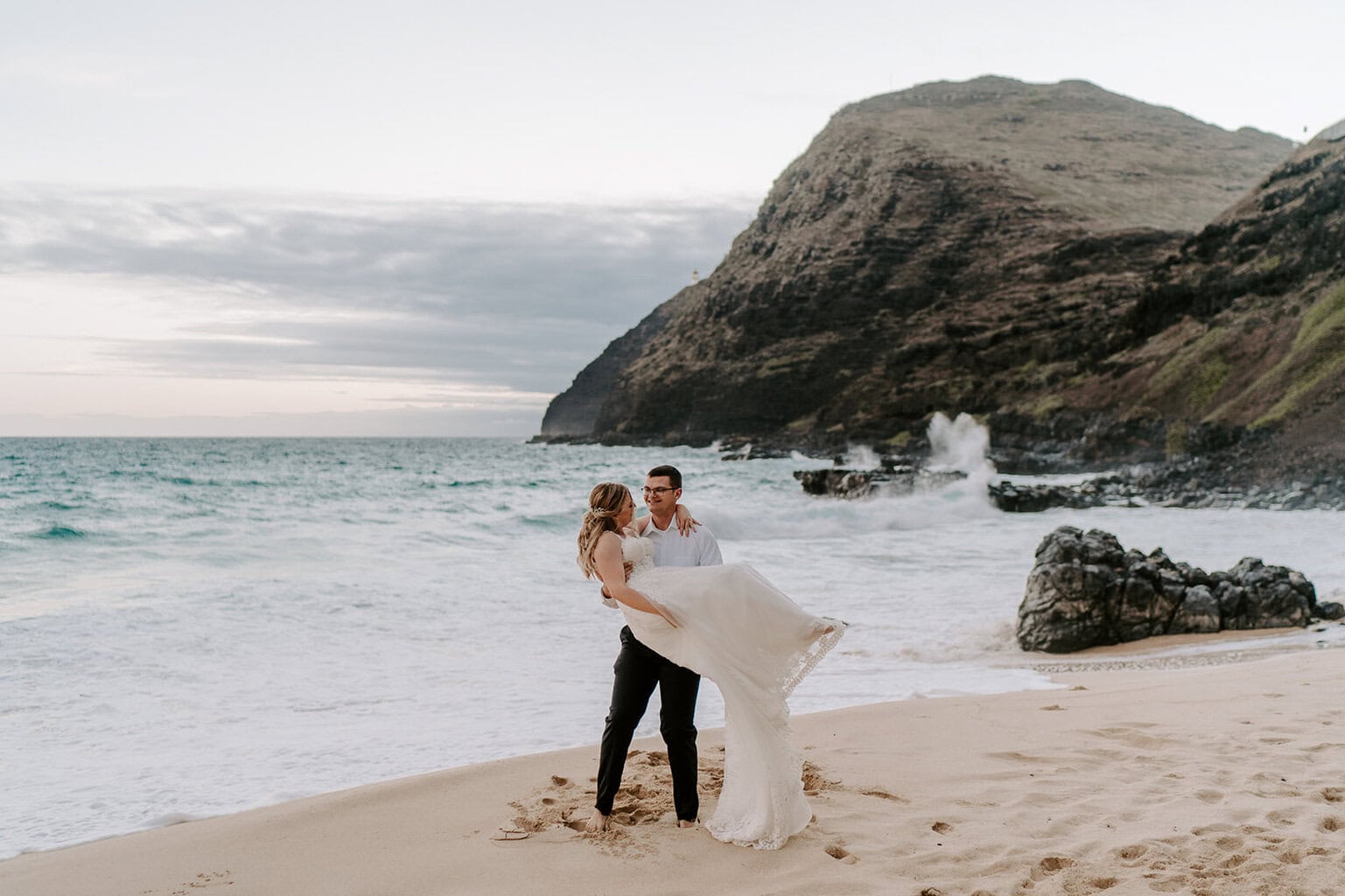 newlyweds walking along the beaches of oahu with a mountain in the background.