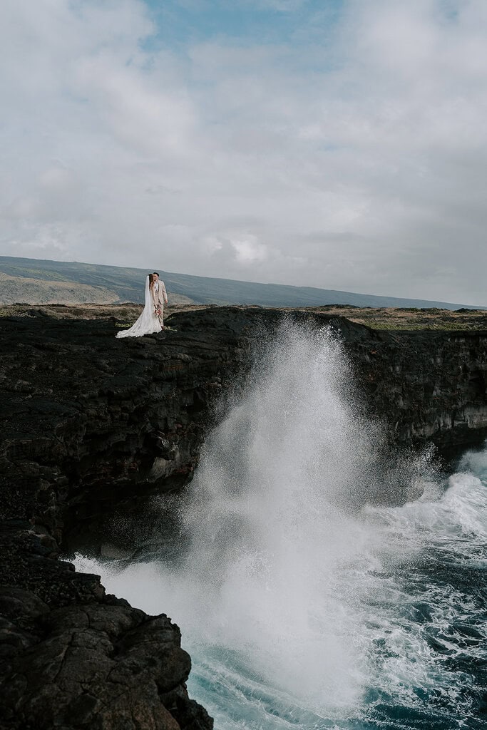 newlyweds hugging as a large wave crashes above a cliffside in Hawaii.