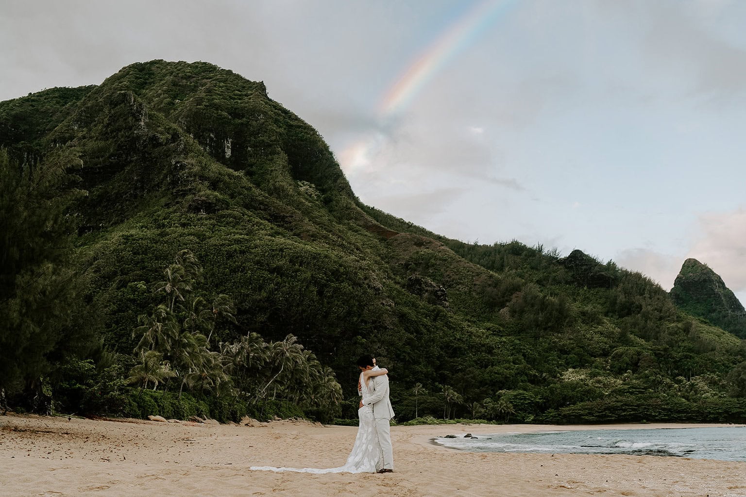 newlyweds kissing on the beach under a rainbow in hawaii.