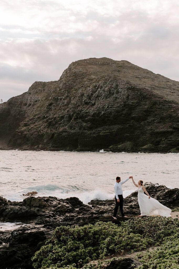 Newlyweds dancing on a beach with a mountain in the background during their Oahu elopement.