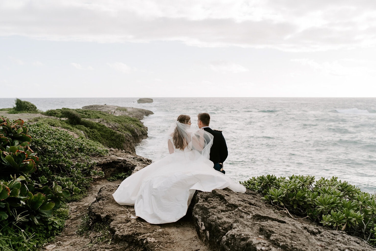 Newlyweds sitting on a cliffside in Hawaii overlooking the ocean.