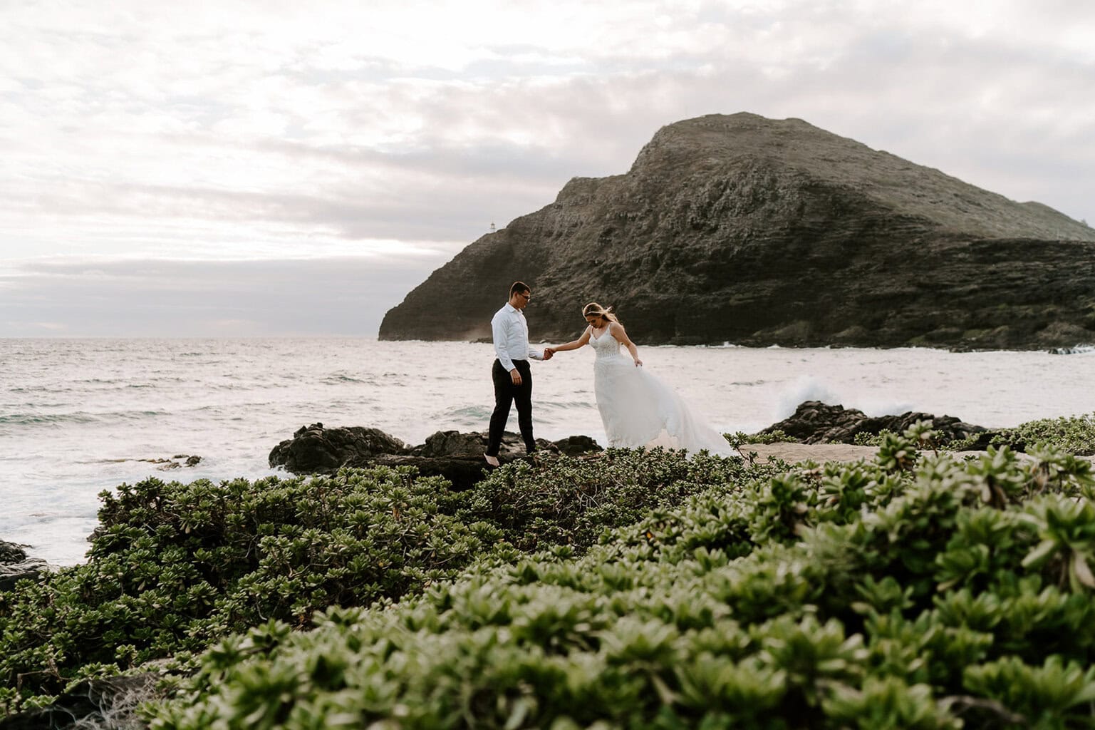 Newlyweds holding hands on the beach with a mountain in the background during their Oahu elopement.
