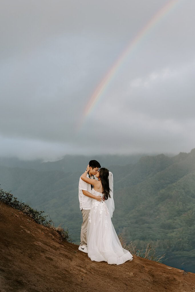 Newlyweds kissing with a rainbow in the background on a cliff in hawaii.