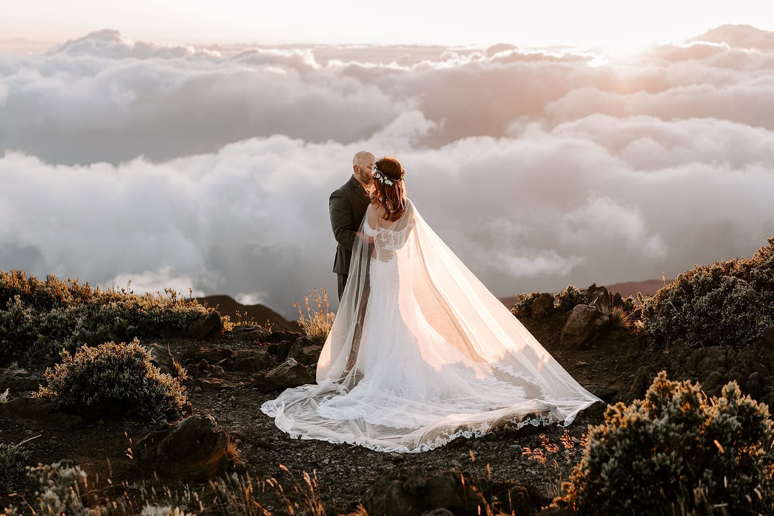 newlyweds holding each other and kissing at sunrise above the clouds in Hawaii. 