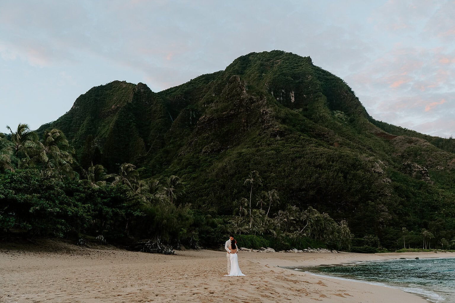 newlyweds hugging on a golden sand beach looking at a towering green mountain during their kauai elopement.