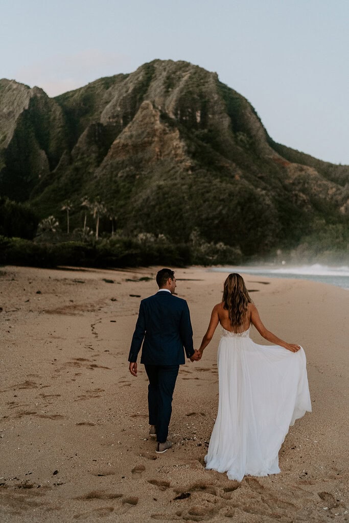 newlyweds holding hands walking on a golden sand beach during their kauai elopement.