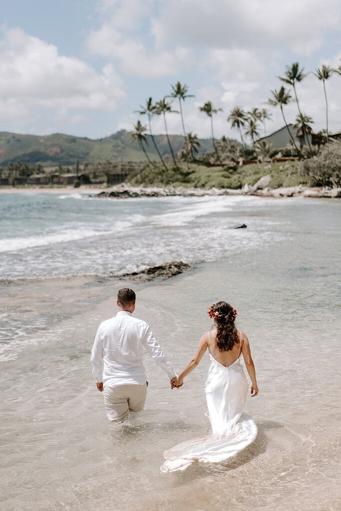 newlyweds holding hands wading into the water during their trash the dress hawaii session.