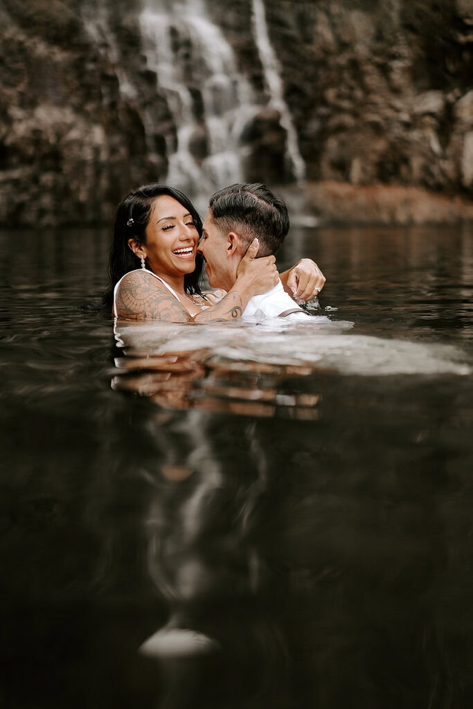 newlyweds wading in a pool of water in front of a waterfall.