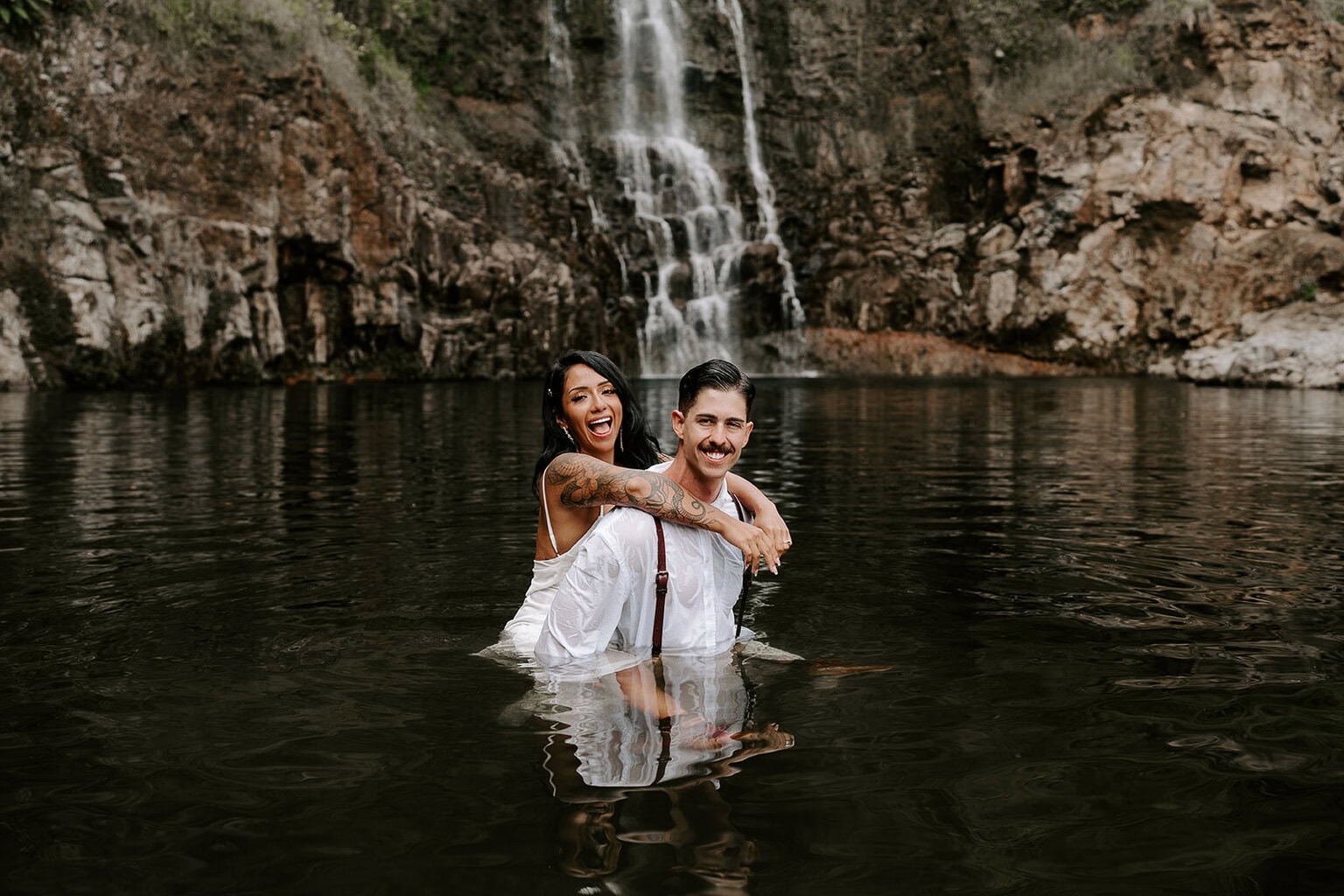 Newlyweds smiling wading by a waterfall during their trash the dress Hawaii session.