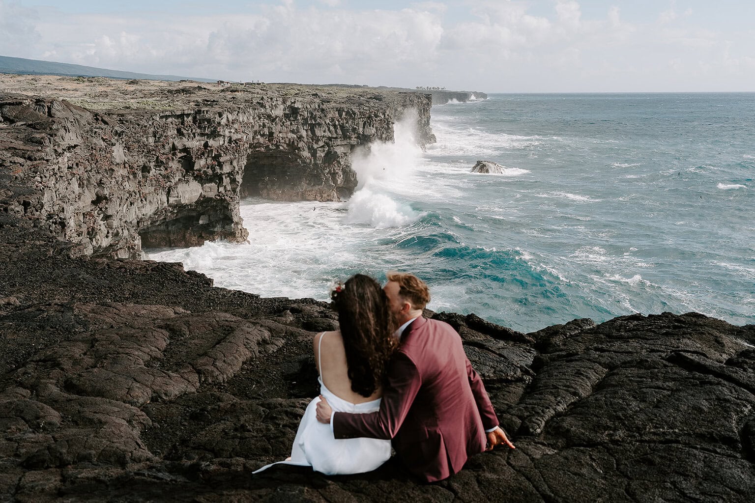 newlyweds sitting at the edge of an ocean cliff hugging.