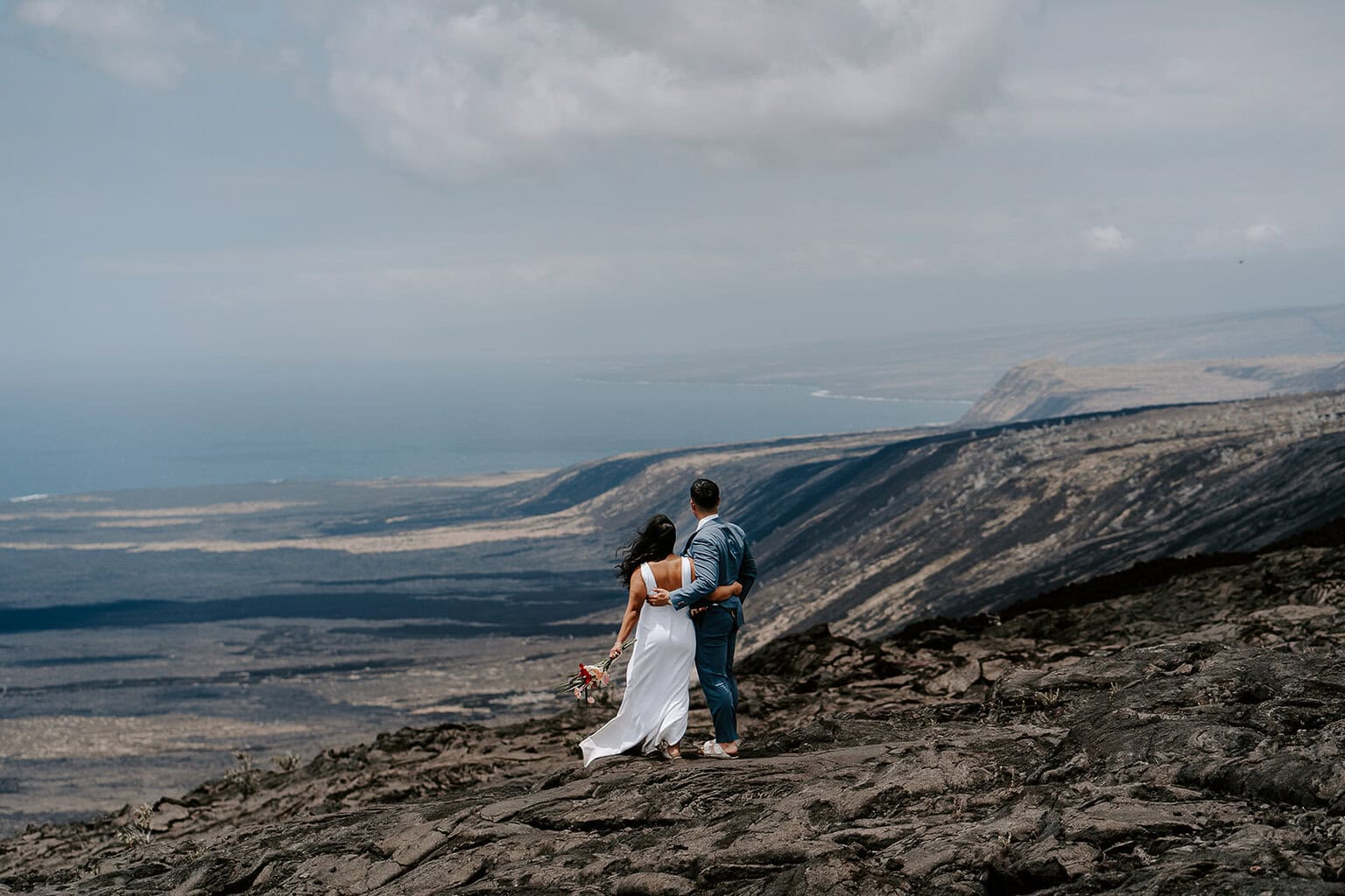 Newlyweds soaking in the view of the ocean standing on a lava field.