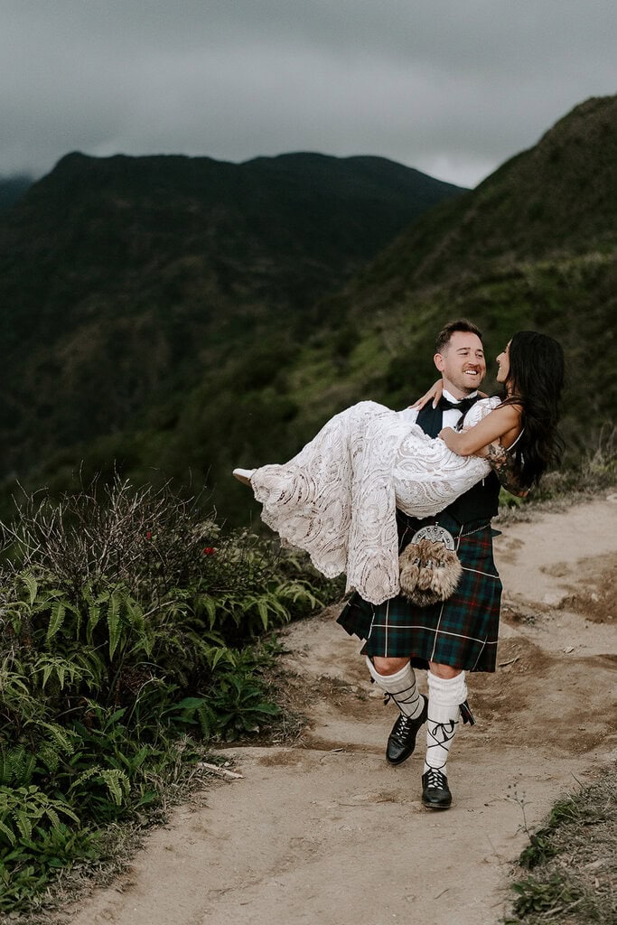 the husband carrying his wife as they hike along a trail through green mountains, one of the best Maui elopement locations.