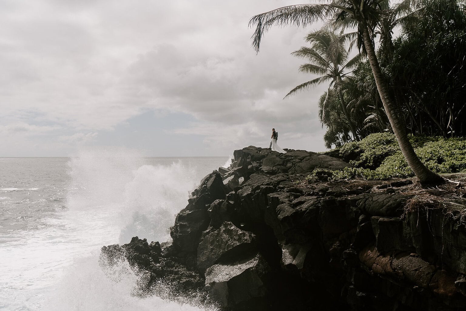 newlyweds standing at the edge of a cliff as waves crash by them.