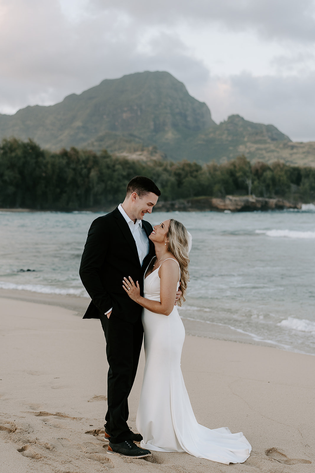 Newlyweds hugging on a beach with a mountain in the background, one of the best Kauai elopement locations.