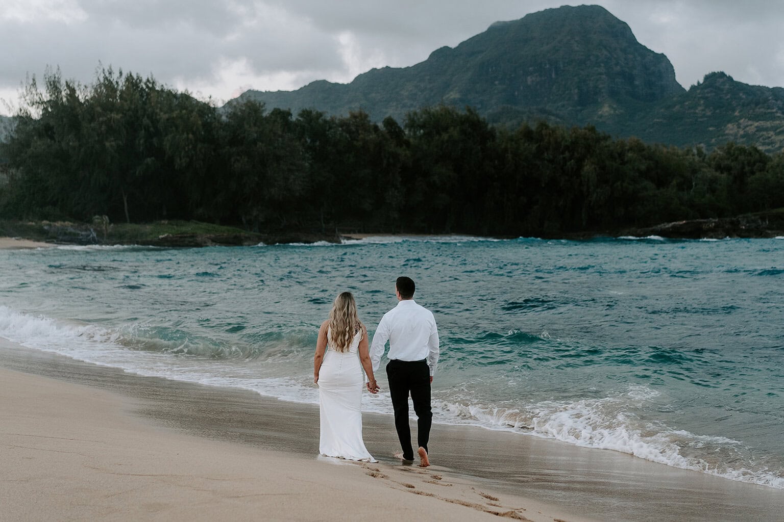 Newlyweds walking along a beach with a green mountain in the background, one of the best Kauai elopement locations.