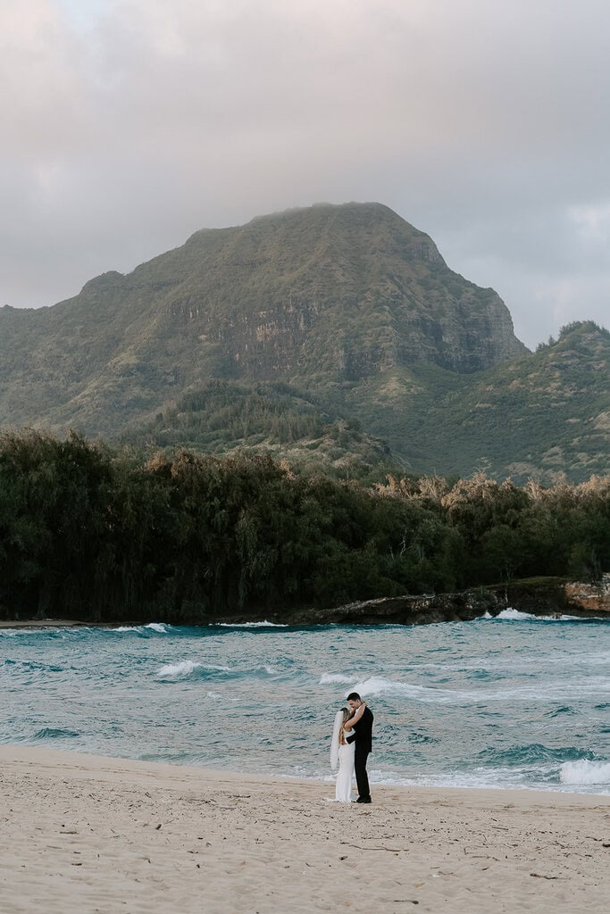 Newlyweds hugging on a beach with a green mountain in the backgrond, one of the best Kauai elopement locations.