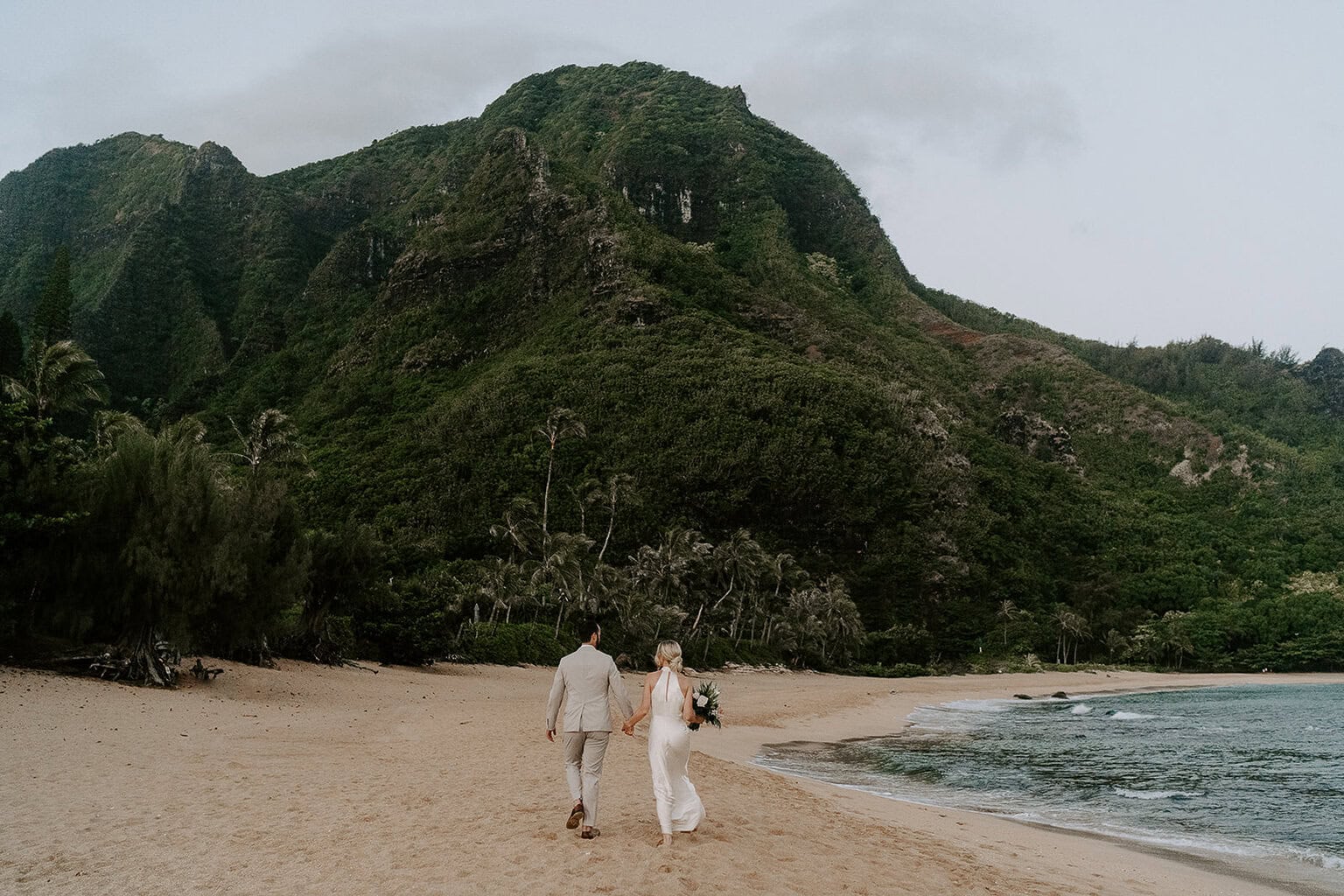 Newlyweds holding hands walking along a beach in Kauai.