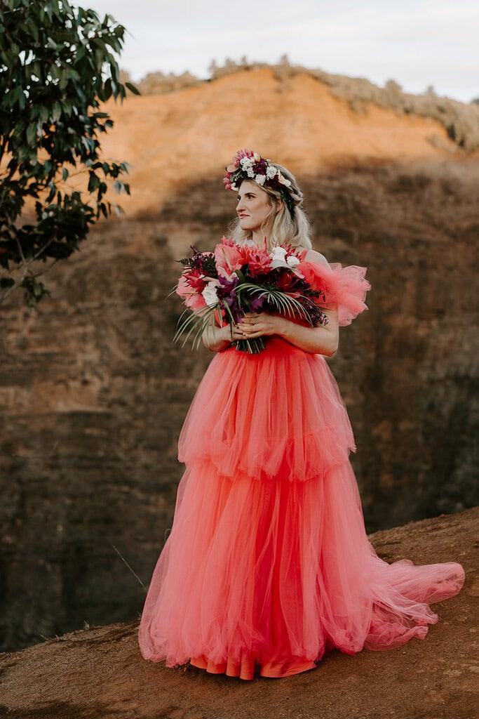 A bride in a pink dress smiles and looks off at the distance while the rust colored canyon wall behind her is lit by the golden hour sun.