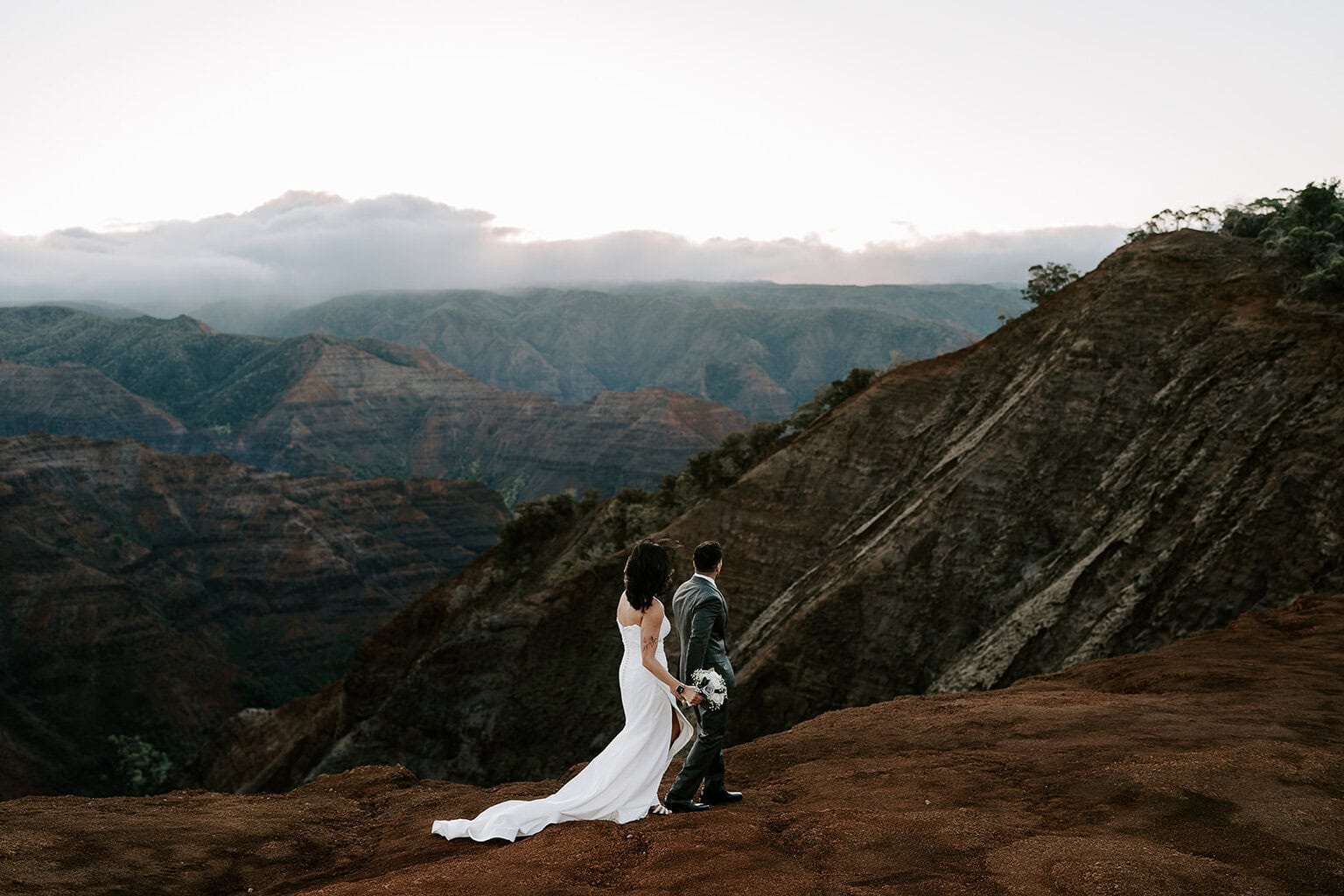 newlyweds hiking along the rim of waimea canyon in their wedding attire.